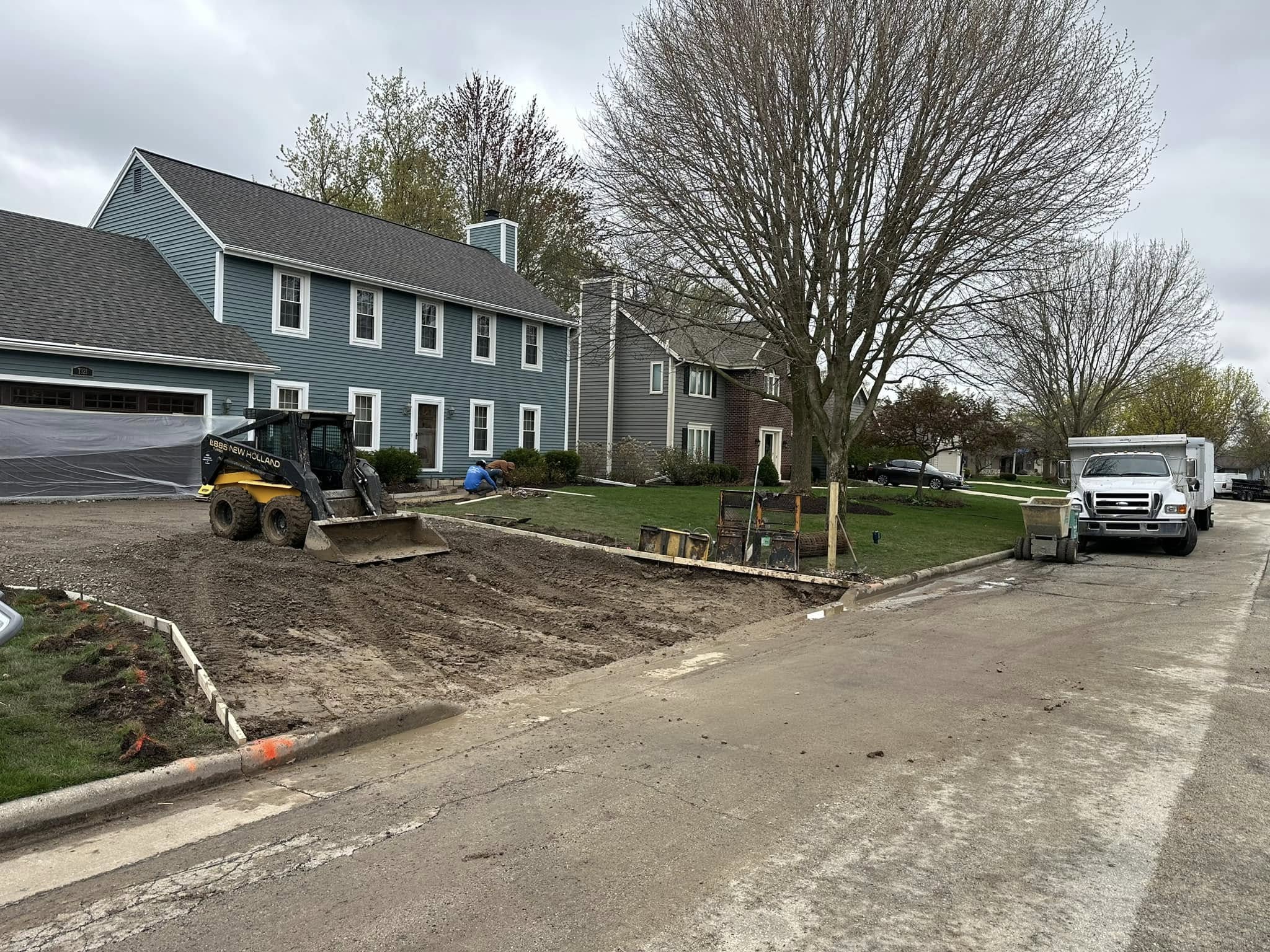 Skid steer excavating and clearing existing driveway area in front of blue colonial home at start of demolition